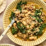 A bowl of Sausage and White Bean Soup with leafy kale in a savory broth, garnished with red pepper flakes. A spoon rests in the bowl, with a small dish of spices nearby.