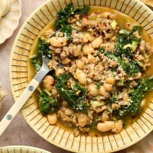 A bowl of Sausage and White Bean Soup with leafy kale in a savory broth, garnished with red pepper flakes. A spoon rests in the bowl, with a small dish of spices nearby.