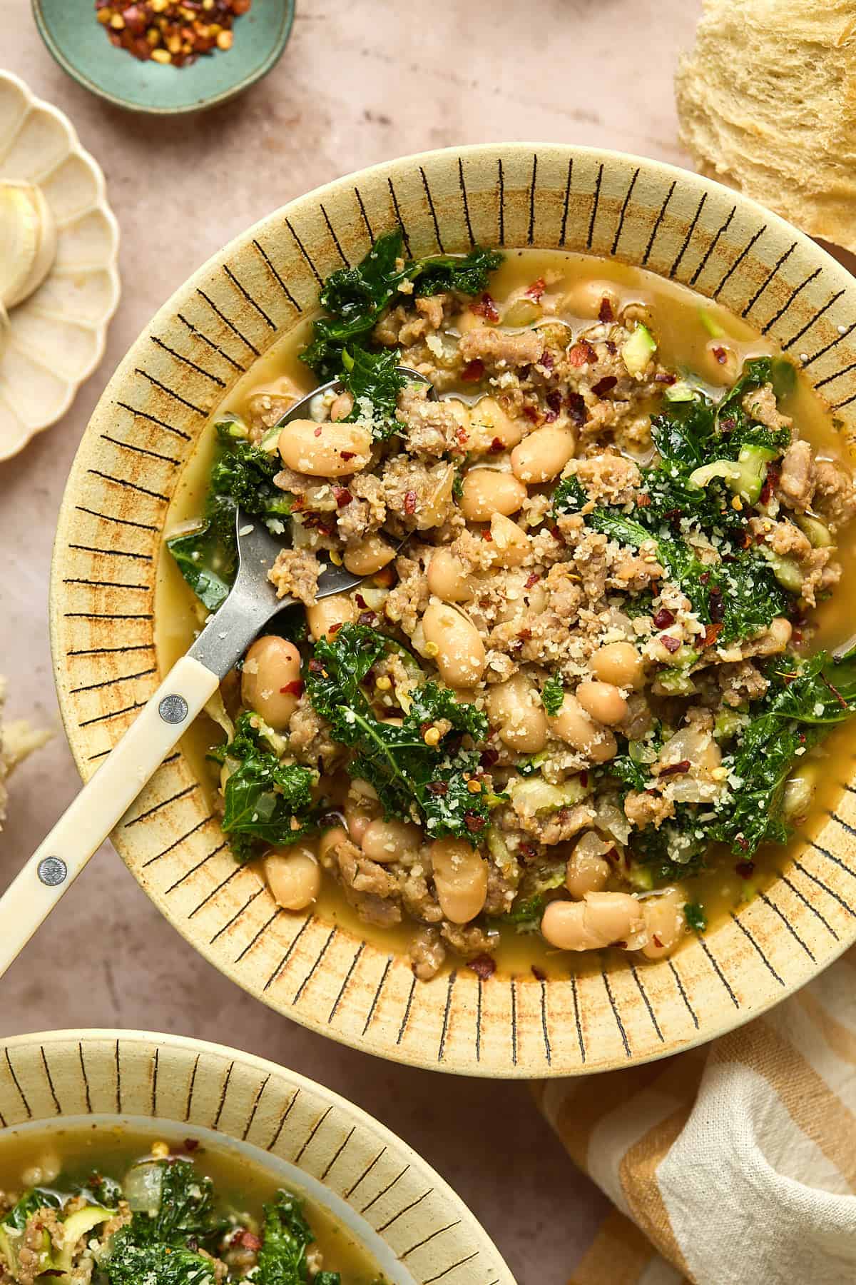 A bowl of Sausage and White Bean Soup with leafy kale in a savory broth, garnished with red pepper flakes. A spoon rests in the bowl, with a small dish of spices nearby.