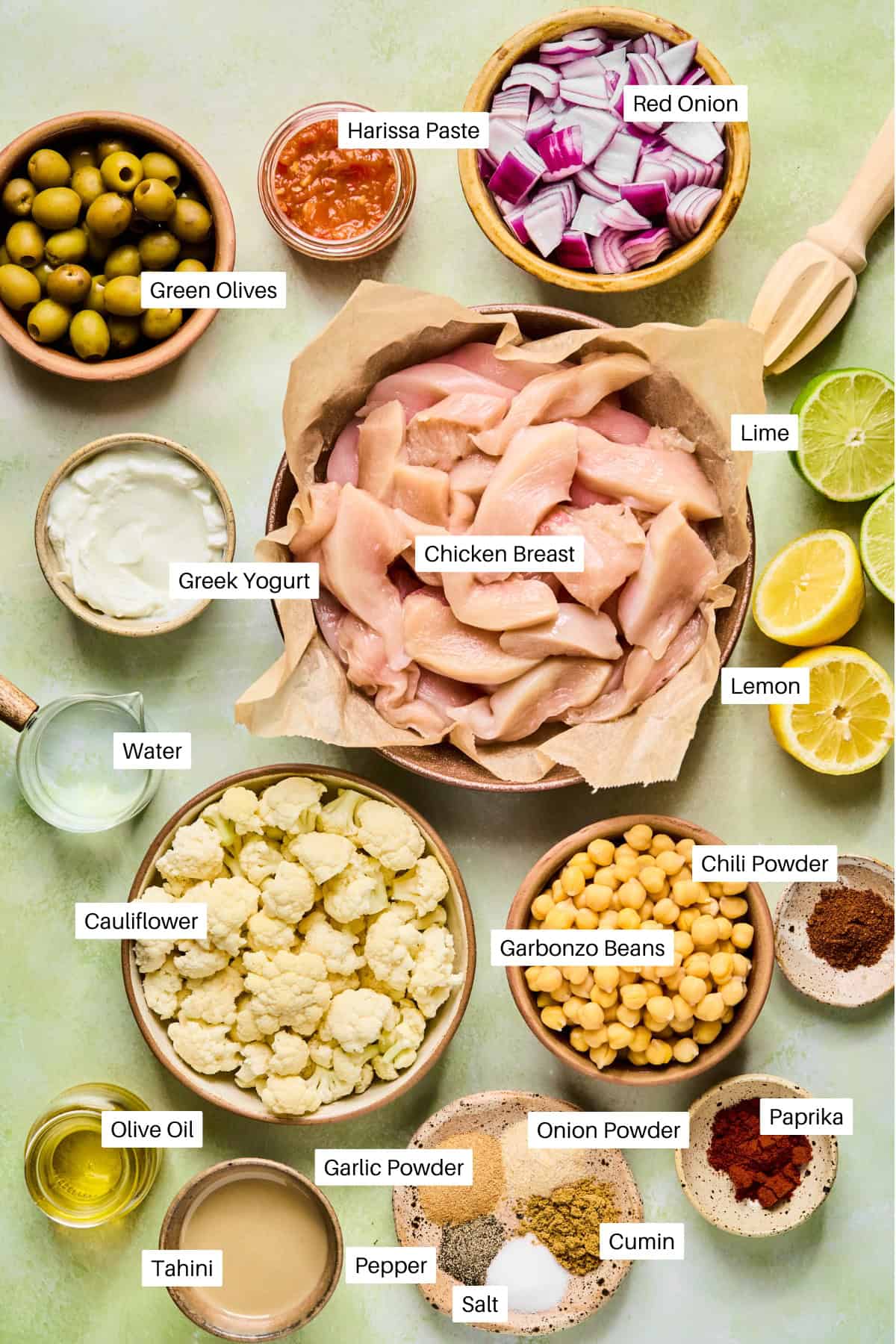 Overhead shot of labeled ingredients for a harissa tray bake chicken recipe, including raw chicken breast, green olives, harissa paste, red onion, citrus, Greek yogurt, cauliflower, garbanzo beans, spices, tahini, olive oil, and water.