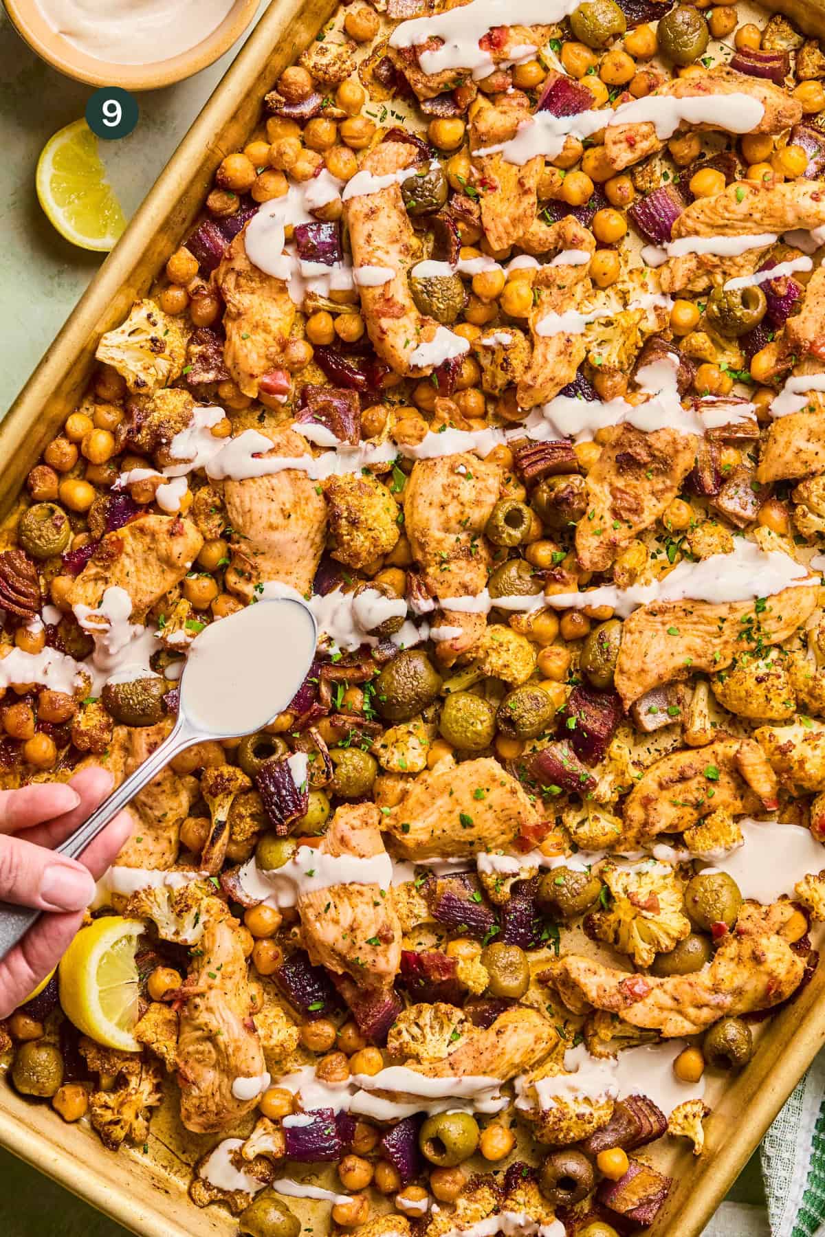 A sheet pan filled with harissa tray bake chicken, chickpeas, olives, red onions, and cauliflower, drizzled with creamy sauce. A hand holds a spoon to add more sauce. Lemon wedges are on the side.