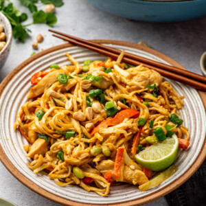 A plate of Chicken Satay Noodles with chicken, vegetables, chopped peanuts, and green onions, garnished with a lime wedge. Chopsticks rest on the plate, and more noodles are in a pan in the background.