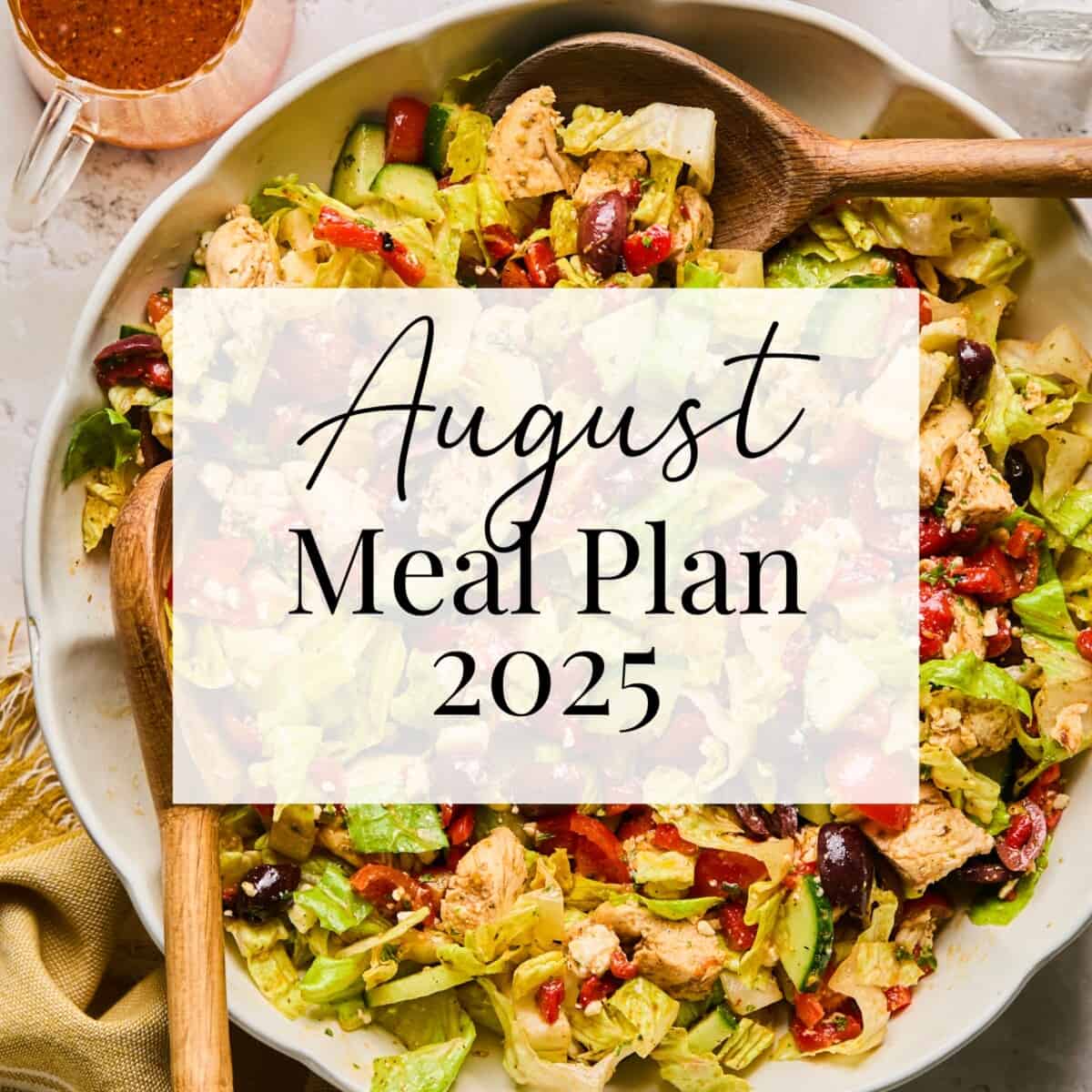 A large salad with lettuce, chicken, olives, and tomatoes in a bowl, with a wooden spoon and small cup of dressing. Text overlay reads August Meal Plan 2025.