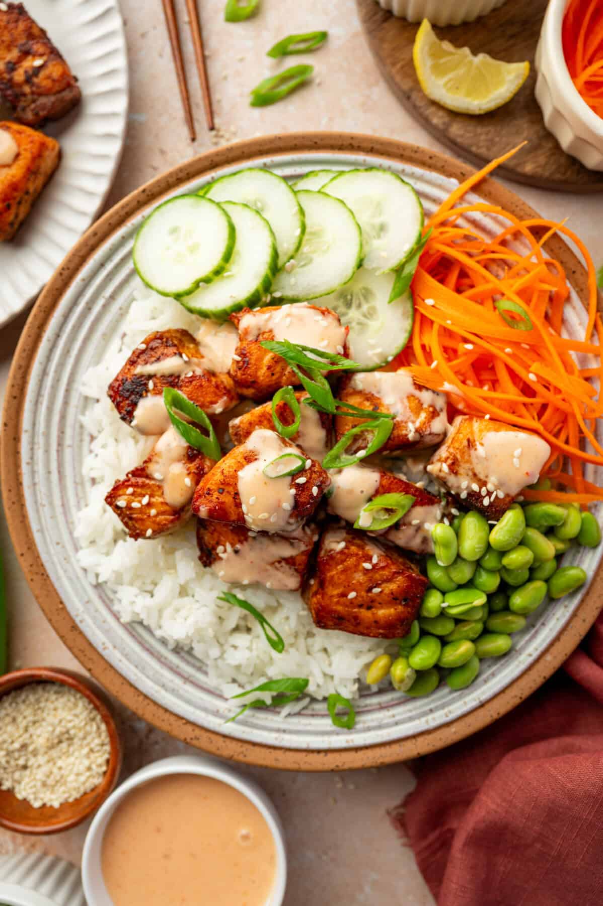 A bowl of white rice topped with seared tofu cubes, sliced cucumbers, shredded carrots, edamame, and a creamy bang bang salmon-inspired sauce, garnished with sliced green onions and served with a lemon wedge.