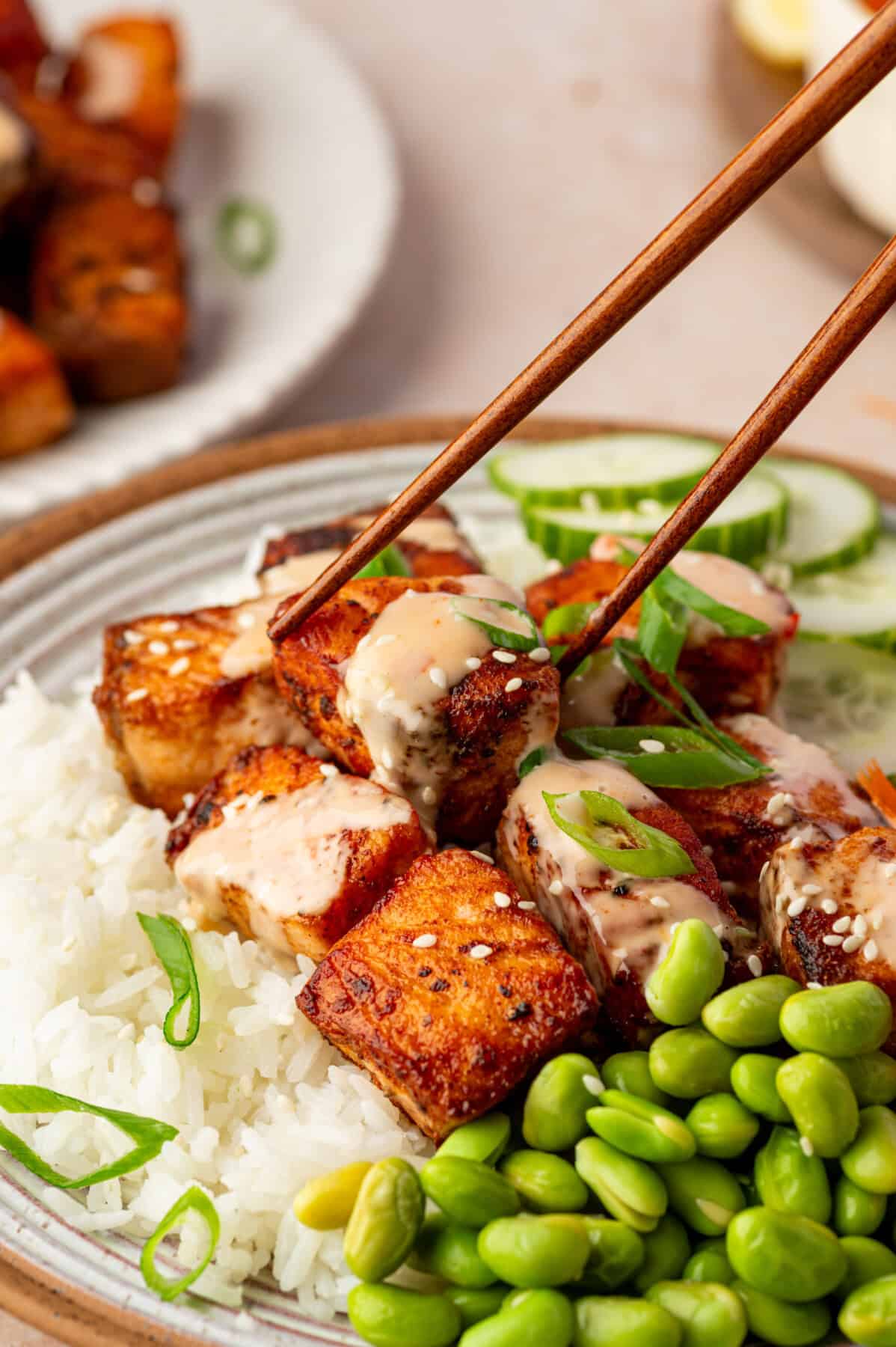A close-up of chopsticks holding a piece of bang bang salmon-glazed tofu over a bowl filled with white rice, edamame, sliced cucumber, and topped with a creamy sauce and green onions.