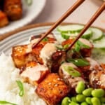 A close-up of chopsticks holding a piece of bang bang salmon-glazed tofu over a bowl filled with white rice, edamame, sliced cucumber, and topped with a creamy sauce and green onions.