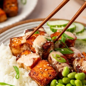 A close-up of chopsticks holding a piece of bang bang salmon-glazed tofu over a bowl filled with white rice, edamame, sliced cucumber, and topped with a creamy sauce and green onions.
