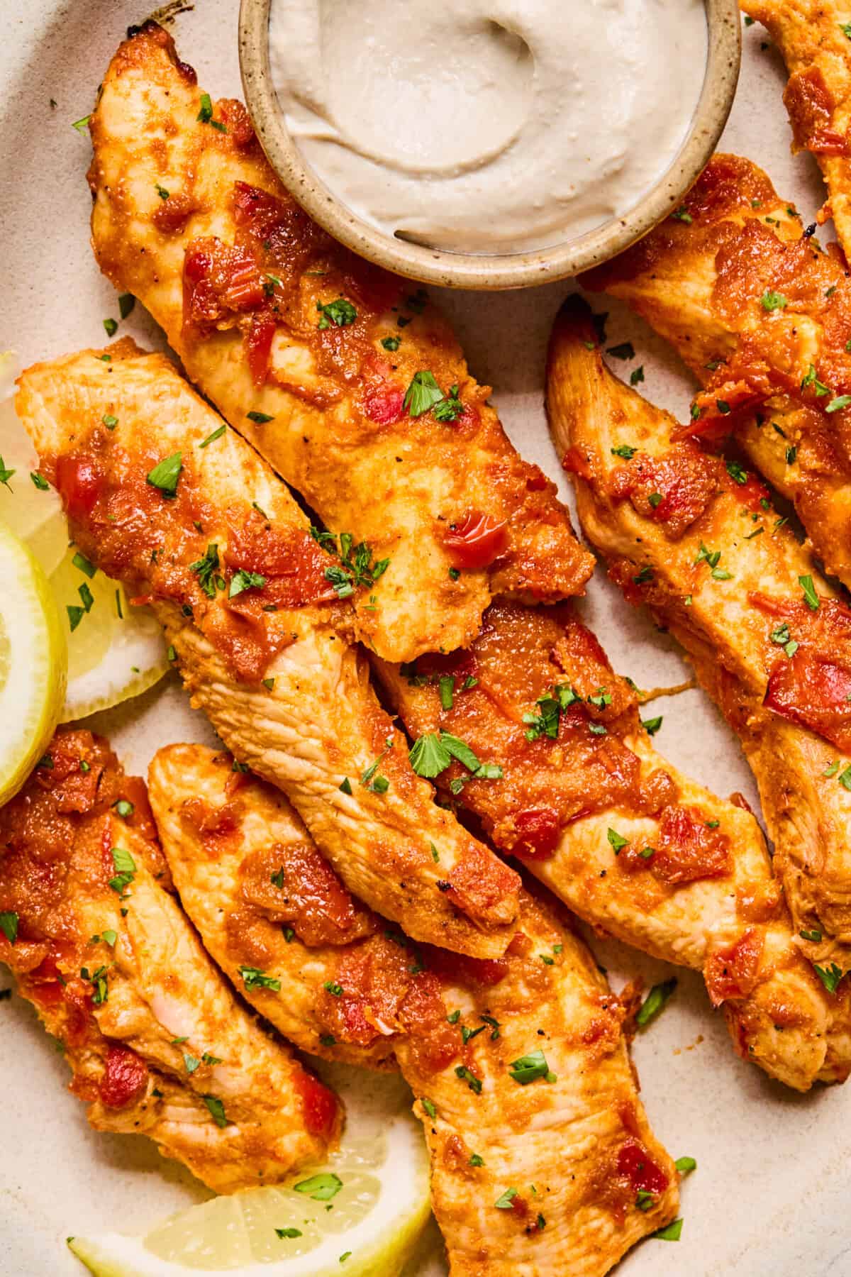 Grilled harissa chicken strips topped with a tomato-based sauce, garnished with chopped herbs, served with lemon wedges and a small bowl of creamy dipping sauce on a beige plate.