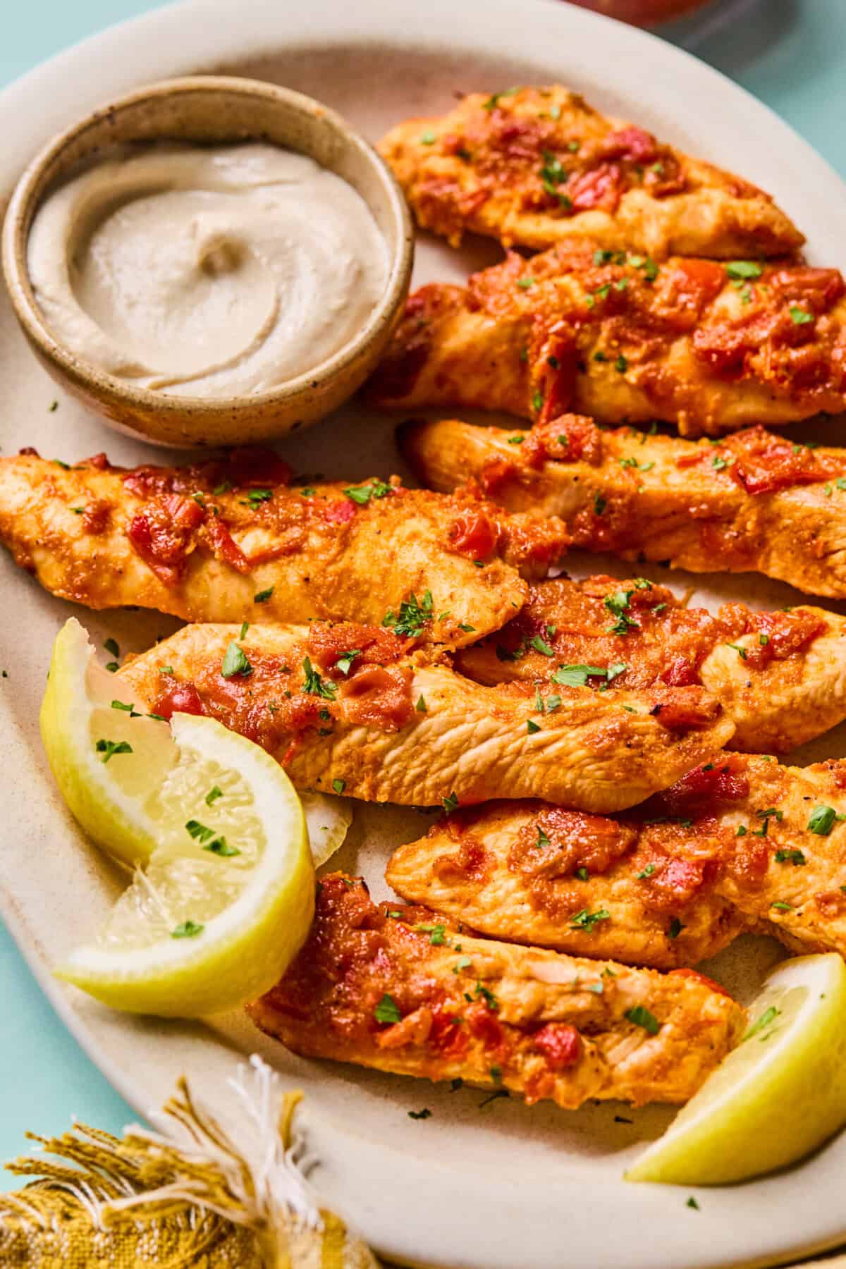 Grilled harissa chicken strips topped with herbs and tomato sauce, served with lemon wedges and a small bowl of creamy dipping sauce on an oval plate.