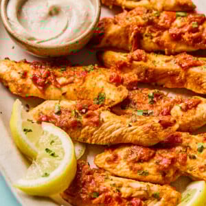 Grilled harissa chicken strips topped with herbs and tomato sauce, served with lemon wedges and a small bowl of creamy dipping sauce on an oval plate.