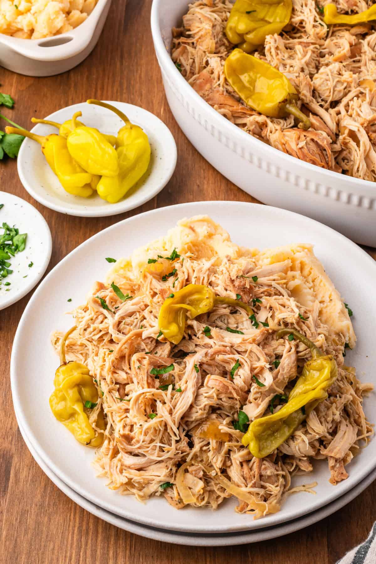 A plate of Mississippi Slow Cooker Chicken topped with sliced pepperoncini peppers is served alongside a casserole dish filled with more tender chicken and peppers, all set on a wooden table.