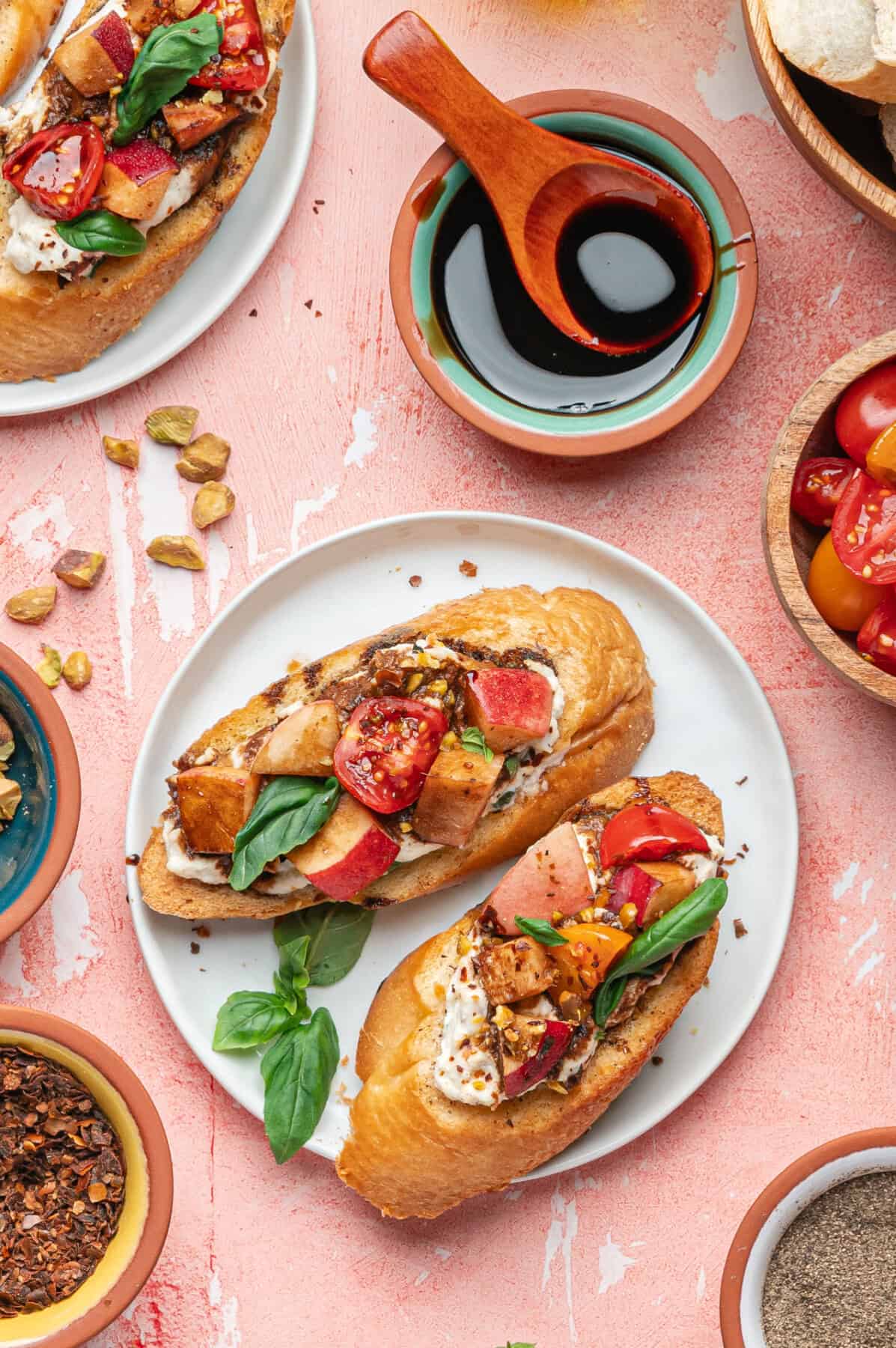 Two pieces of toasted bread topped with creamy cheese, grilled chicken, tomatoes, basil, and pistachios create a delicious ricotta bruschetta, served on a white plate surrounded by bowls of tomatoes, spices, nuts, and a dish of dark sauce.