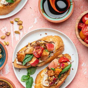 Two pieces of toasted bread topped with creamy cheese, grilled chicken, tomatoes, basil, and pistachios create a delicious ricotta bruschetta, served on a white plate surrounded by bowls of tomatoes, spices, nuts, and a dish of dark sauce.