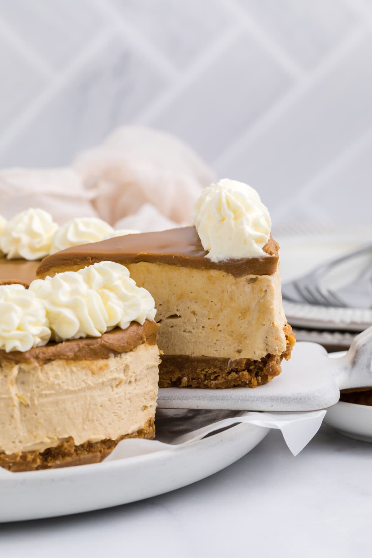 A slice of creamy no bake Biscoff cheesecake with a graham cracker crust and caramel topping, garnished with swirls of whipped cream, is lifted from a white plate. A blurred background shows more cheesecake, napkins, and stacked utensils.