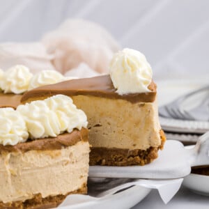A slice of creamy no bake Biscoff cheesecake with a graham cracker crust and caramel topping, garnished with swirls of whipped cream, is lifted from a white plate. A blurred background shows more cheesecake, napkins, and stacked utensils.