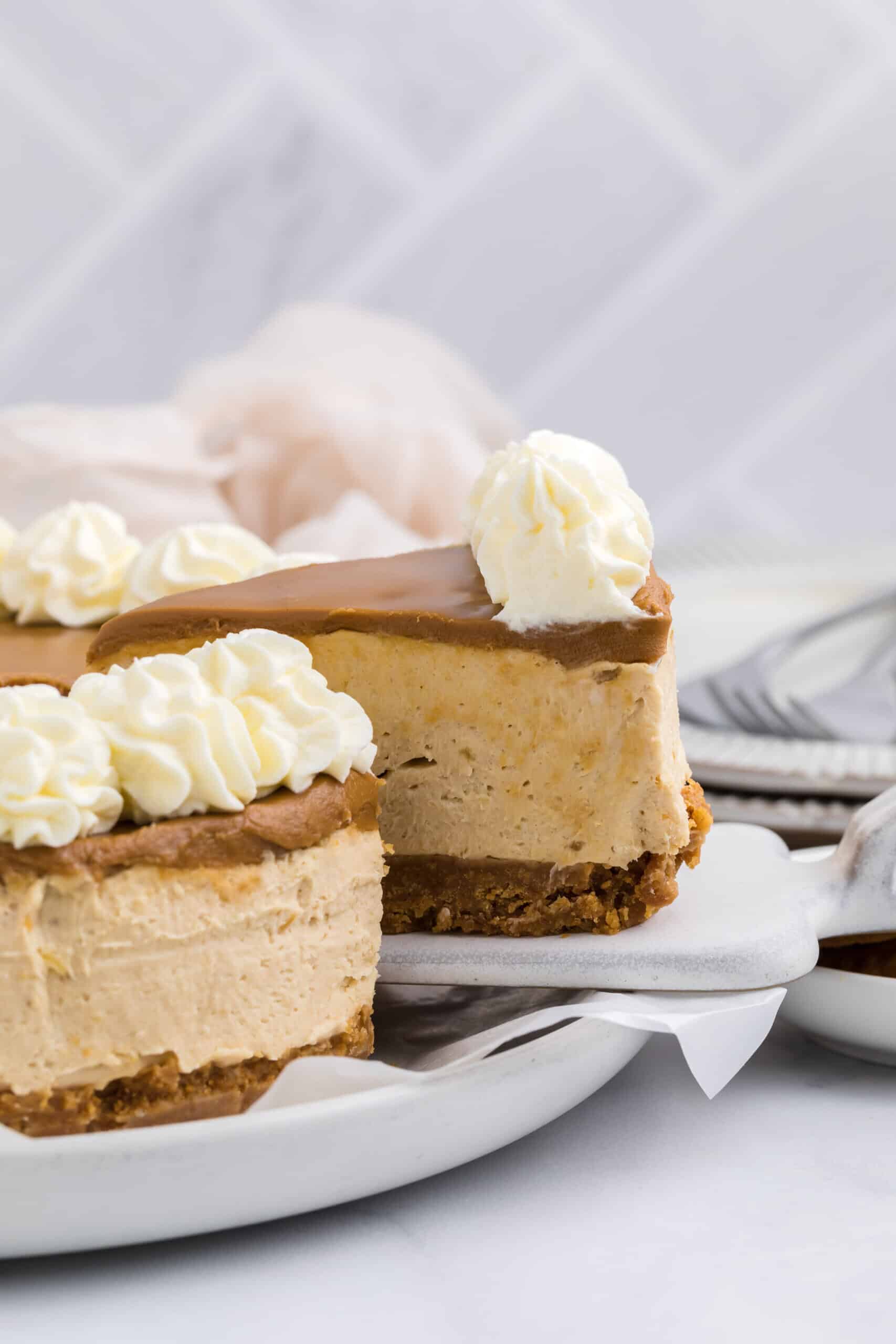 A slice of creamy no bake Biscoff cheesecake with a graham cracker crust and caramel topping, garnished with swirls of whipped cream, is lifted from a white plate. A blurred background shows more cheesecake, napkins, and stacked utensils.