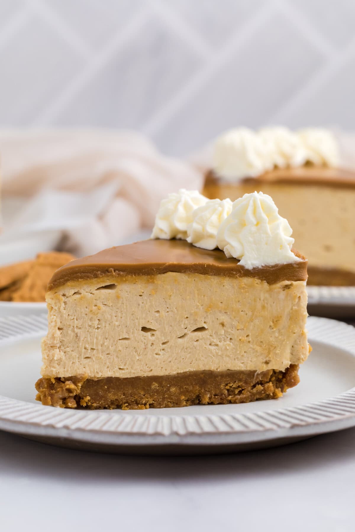 A slice of creamy no bake biscoff cheesecake with a graham cracker crust, topped with caramel sauce and whipped cream, sits on a white plate with another slice in the background.
