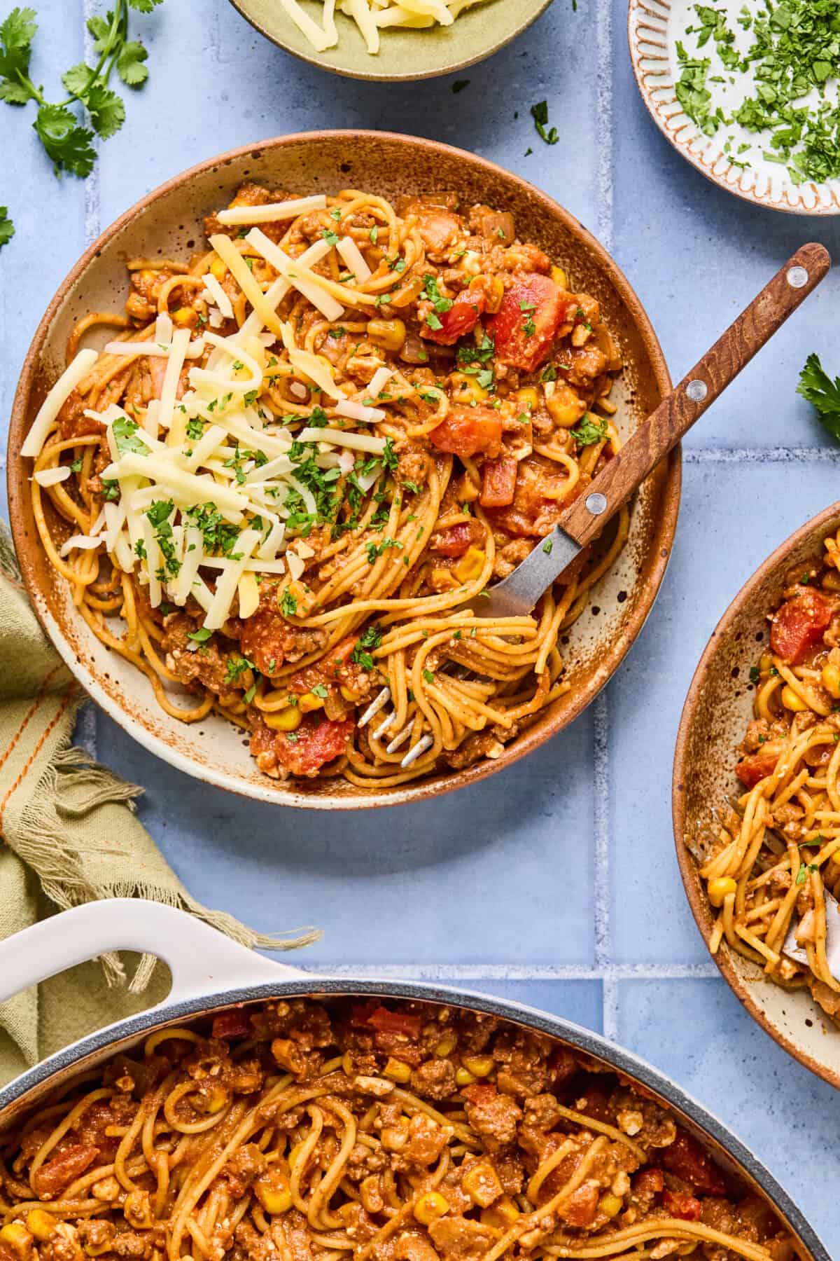A bowl of taco spaghetti mixed with ground meat, tomatoes, corn, and spices, topped with shredded cheese and chopped herbs, sits on a table with a fork. Surrounding bowls hold extra cheese and herbs.