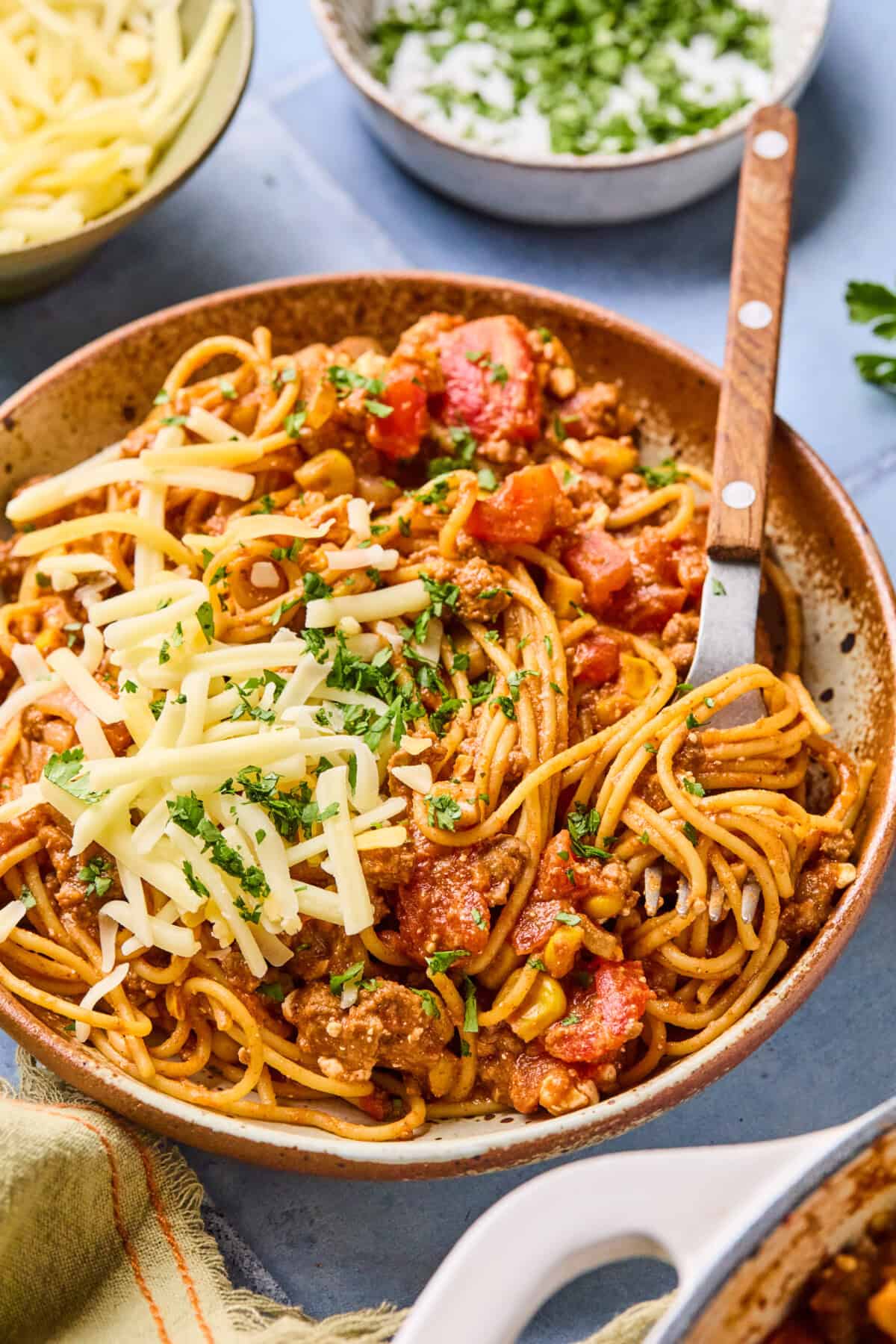 A bowl of taco spaghetti with meat sauce, topped with shredded cheese and chopped herbs, sits on a table with a fork. Nearby are bowls of grated cheese and extra herbs for garnish.