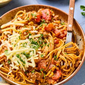 A bowl of taco spaghetti with meat sauce, topped with shredded cheese and chopped herbs, sits on a table with a fork. Nearby are bowls of grated cheese and extra herbs for garnish.