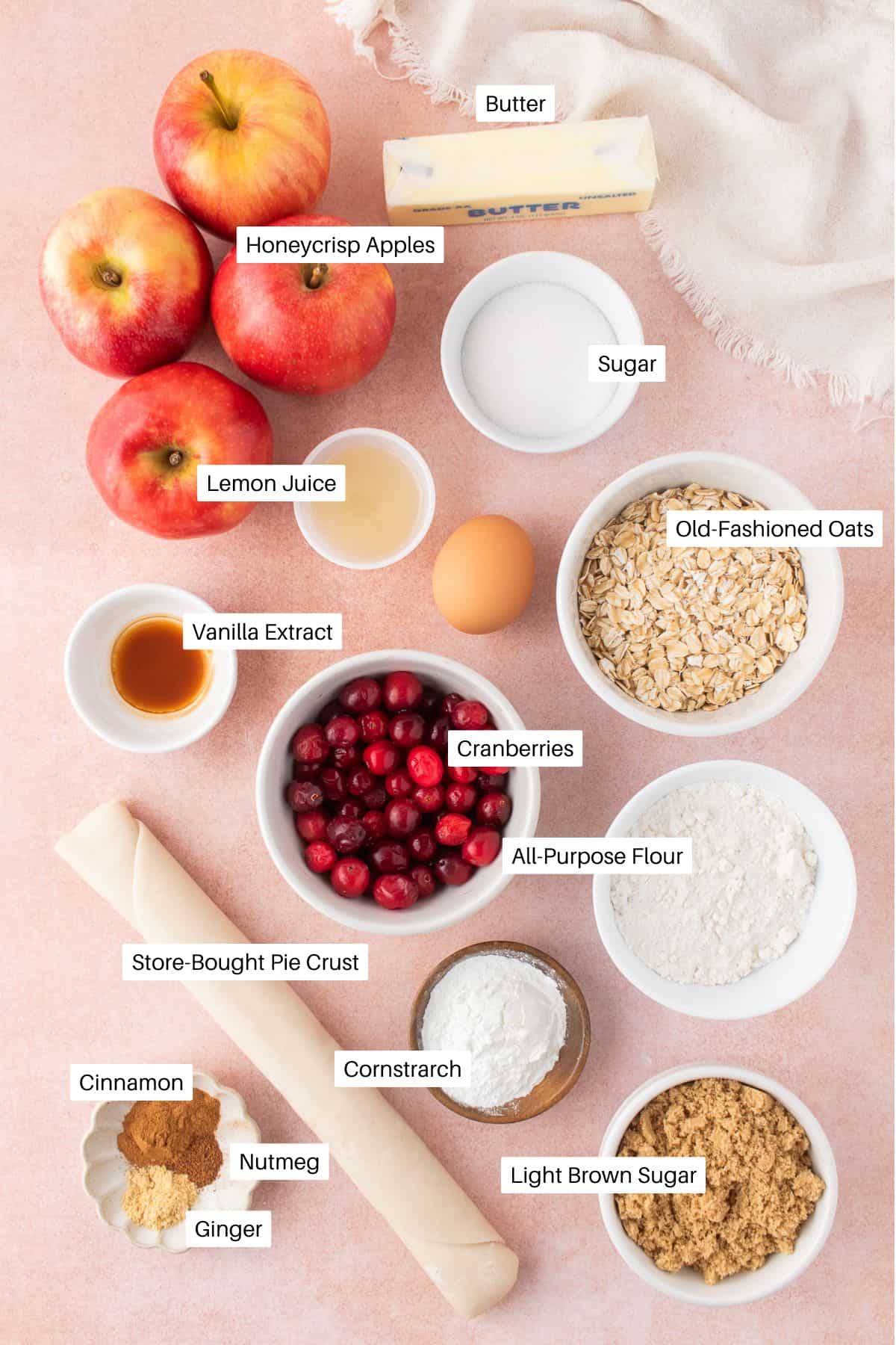 Top-down view of ingredients for a cranberry and apple crumble pie, including apples, cranberries, oats, sugars, spices, lemon juice, egg, butter, vanilla, flour, cornstarch, and a rolled pie crust on a light pink surface with labeled items.