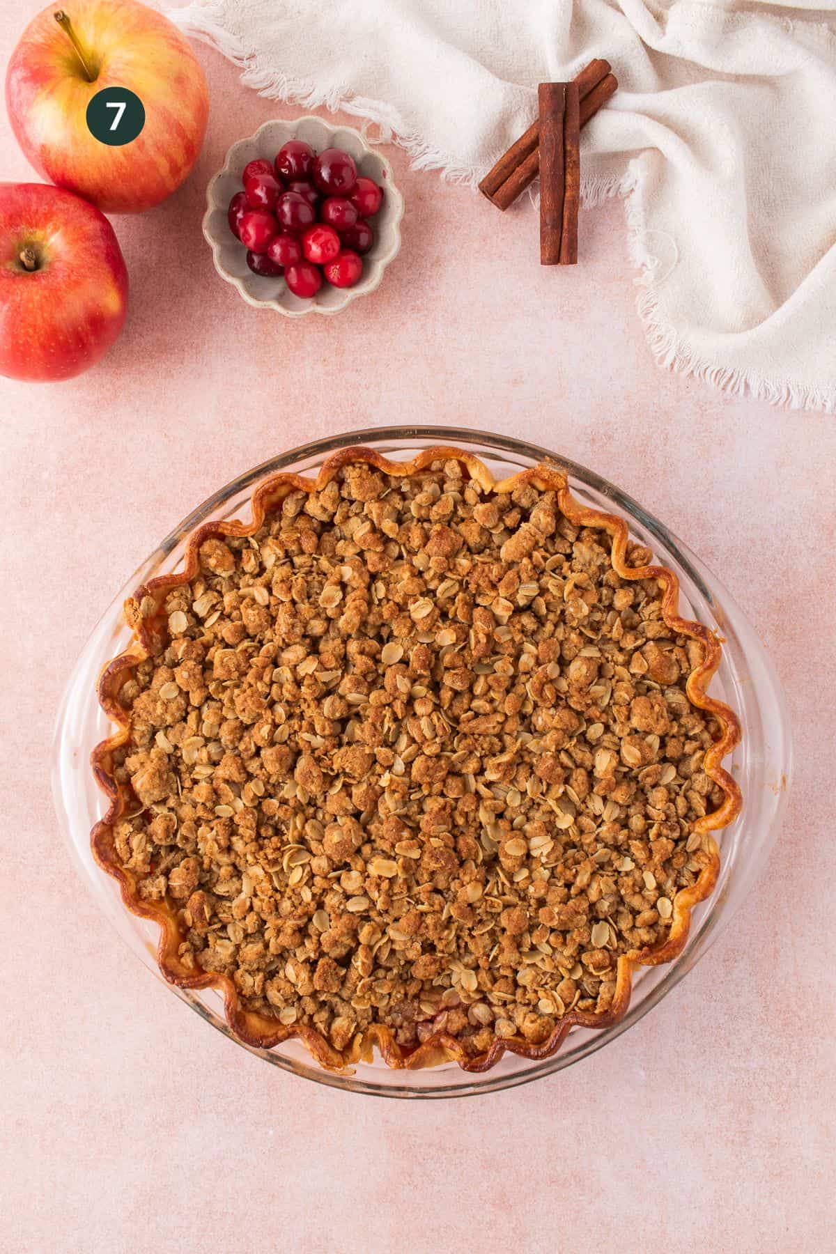 A cranberry and apple crumble pie in a glass dish rests on a light surface, surrounded by two apples, a small bowl of cranberries, cinnamon sticks, and a white cloth.