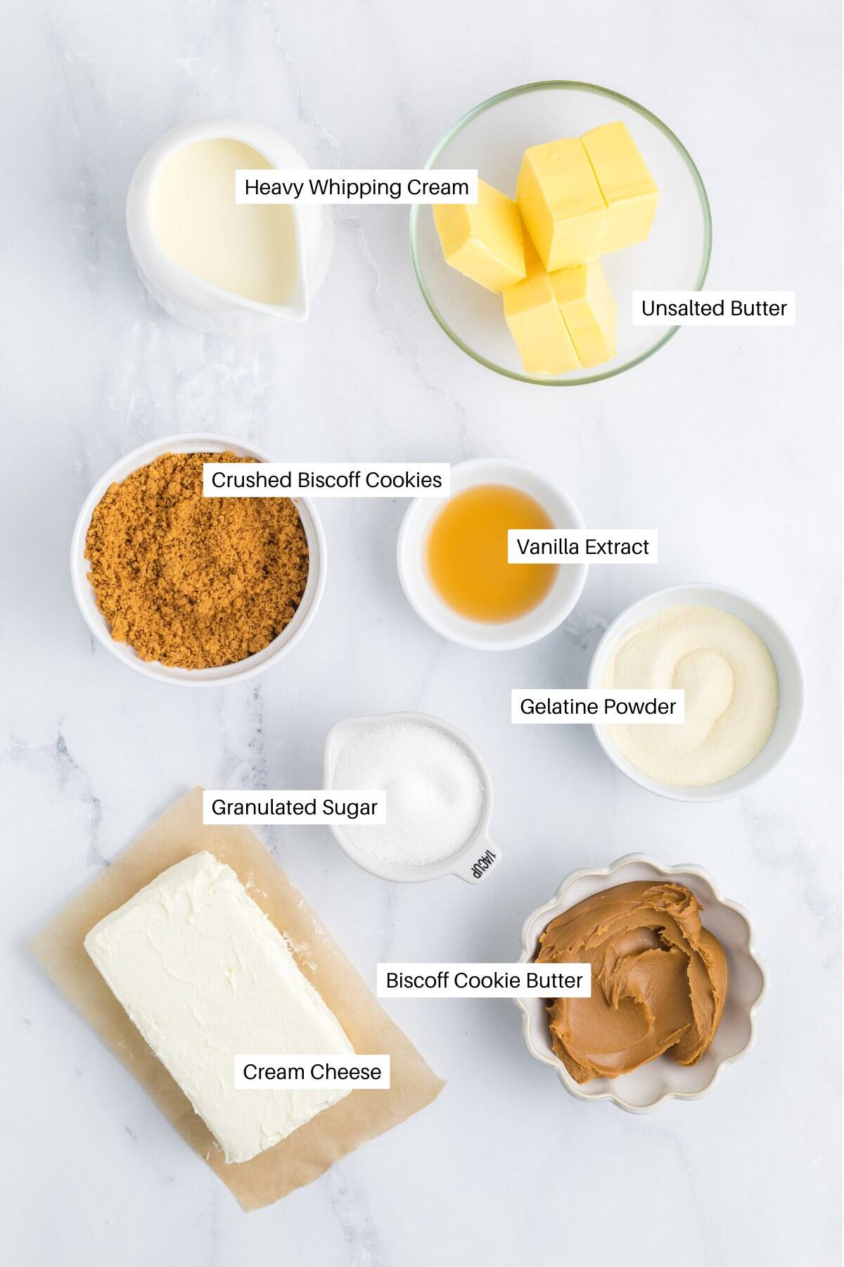 Top-down view of labeled ingredients for no bake Biscoff cheesecake in bowls on a white surface: heavy whipping cream, unsalted butter, crushed Biscoff cookies, vanilla extract, gelatine powder, granulated sugar, Biscoff cookie butter, and cream cheese.
