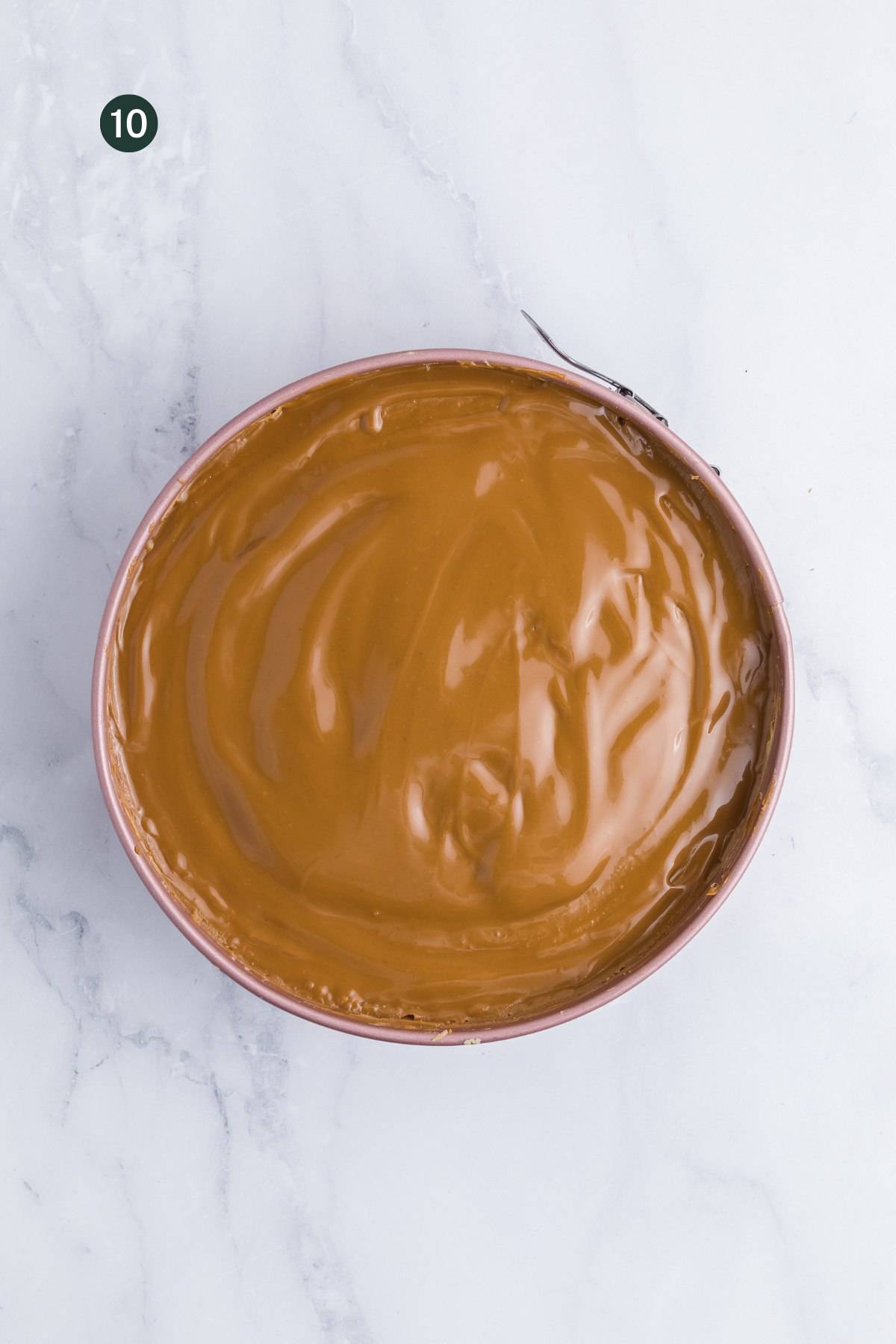 A round cake pan filled with a smooth, shiny layer of no bake biscoff cheesecake topping, viewed from above on a white marble surface. A small black circle with the number 10 is in the top left corner.