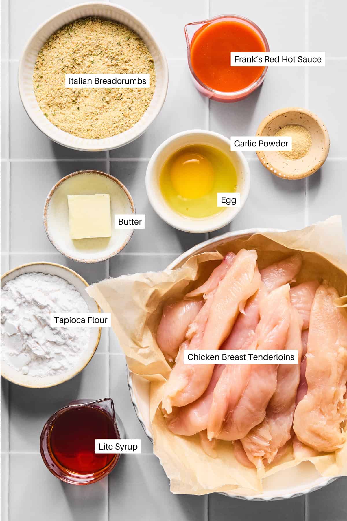 Various ingredients for Air Fryer Buffalo Chicken Tenders are arranged on a white surface, including raw chicken breast tenderloins, Italian breadcrumbs, butter, tapioca flour, lite syrup, an egg, Frank’s Red Hot Sauce, and garlic powder. Labels identify each item.