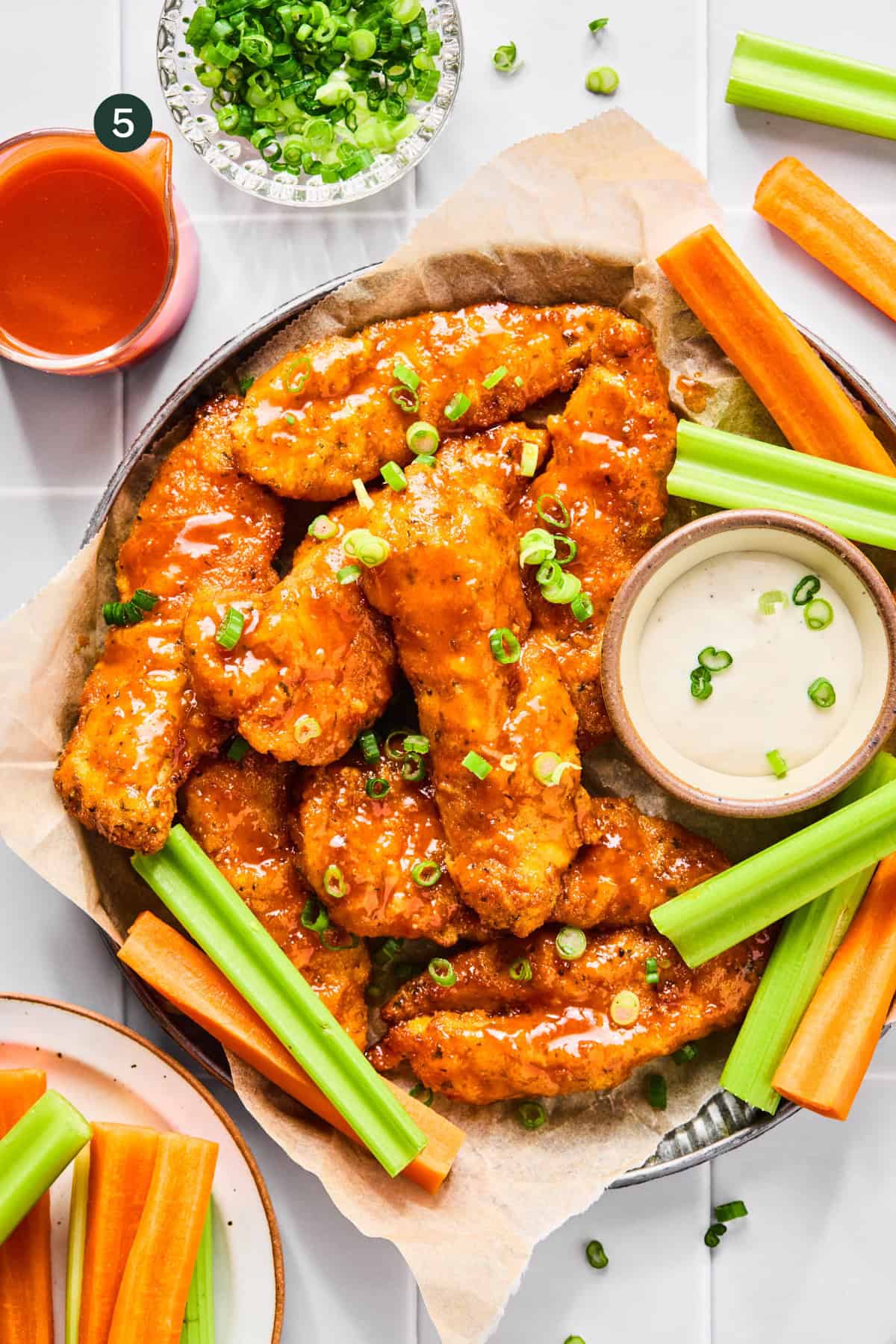 A platter of saucy Air Fryer Buffalo Chicken Tenders garnished with chopped green onions, served with celery and carrot sticks, a bowl of creamy dipping sauce, and a glass of hot sauce on the side.