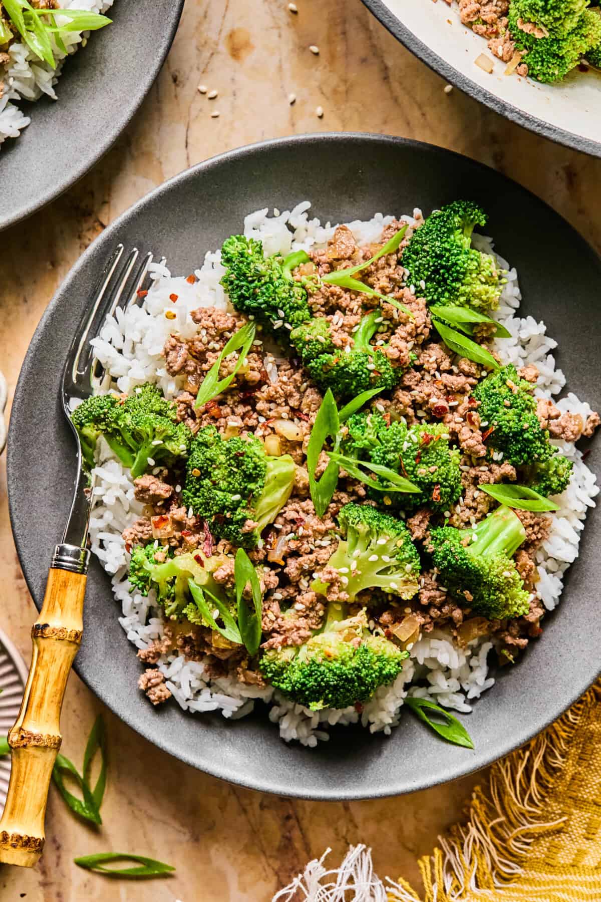 A black bowl filled with white rice is topped with flavorful ground beef and broccoli, garnished with sliced green onions. A bamboo-handled fork rests on the side. The bowl sits on a beige surface near a yellow cloth.
