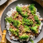 A black bowl filled with white rice is topped with flavorful ground beef and broccoli, garnished with sliced green onions. A bamboo-handled fork rests on the side. The bowl sits on a beige surface near a yellow cloth.
