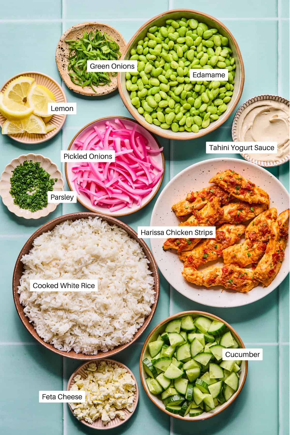 A top view of Harissa Chicken Bowls features labeled ingredients: lemon, green onions, edamame, tahini yogurt sauce, pickled onions, parsley, harissa chicken strips, cooked white rice, cucumber, and feta cheese.