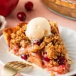A slice of cranberry and apple crumble pie topped with vanilla ice cream sits on a white plate, garnished with cranberries and oats, with a fork and cinnamon stick nearby.