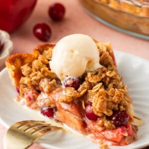 A slice of cranberry and apple crumble pie topped with vanilla ice cream sits on a white plate, garnished with cranberries and oats, with a fork and cinnamon stick nearby.