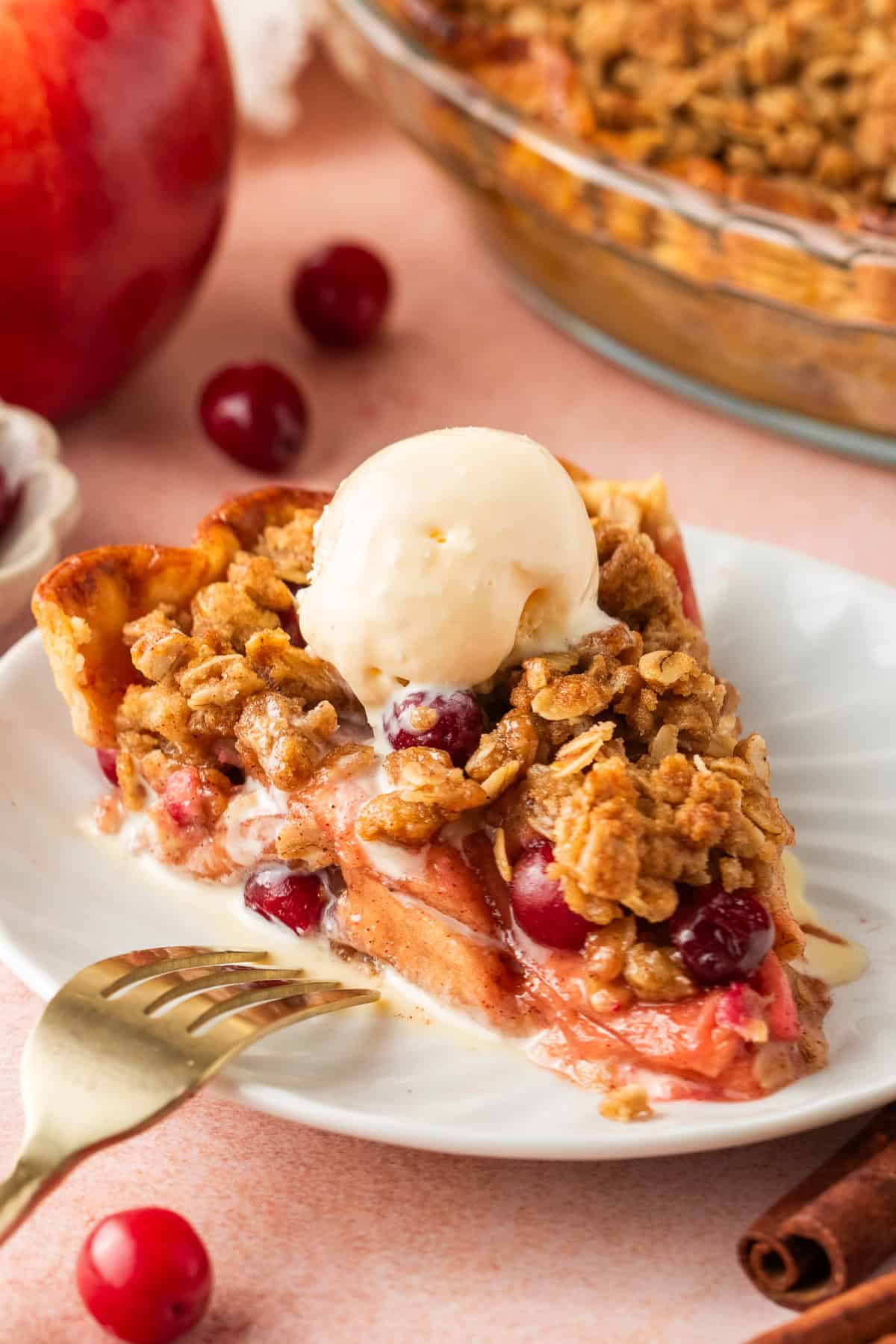 A slice of cranberry and apple crumble pie topped with vanilla ice cream sits on a white plate, garnished with cranberries and oats, with a fork and cinnamon stick nearby.