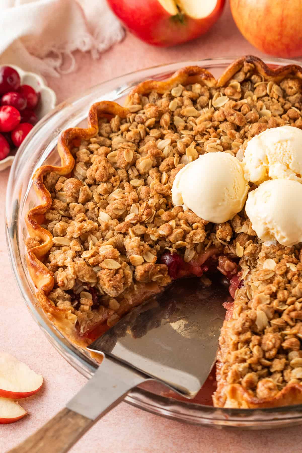 A baked cranberry and apple crumble pie with a golden oat topping and three scoops of vanilla ice cream, with a slice missing. Nearby are fresh apples, cranberries, and a pie server.