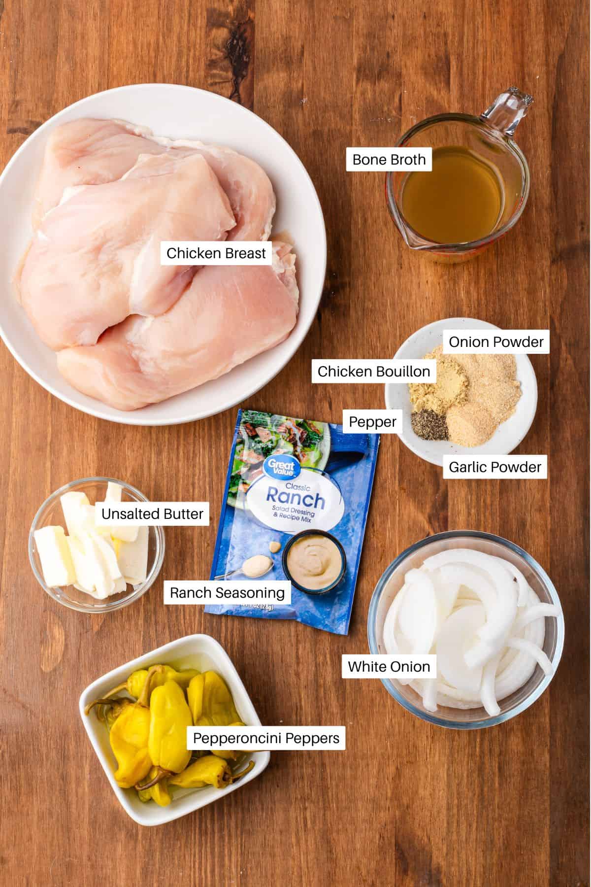 Top-down view of labeled Mississippi Slow Cooker Chicken ingredients—chicken breast, bone broth, unsalted butter, ranch seasoning, pepperoncini peppers, white onion, chicken bouillon, onion powder, pepper, and garlic powder on a wooden surface.