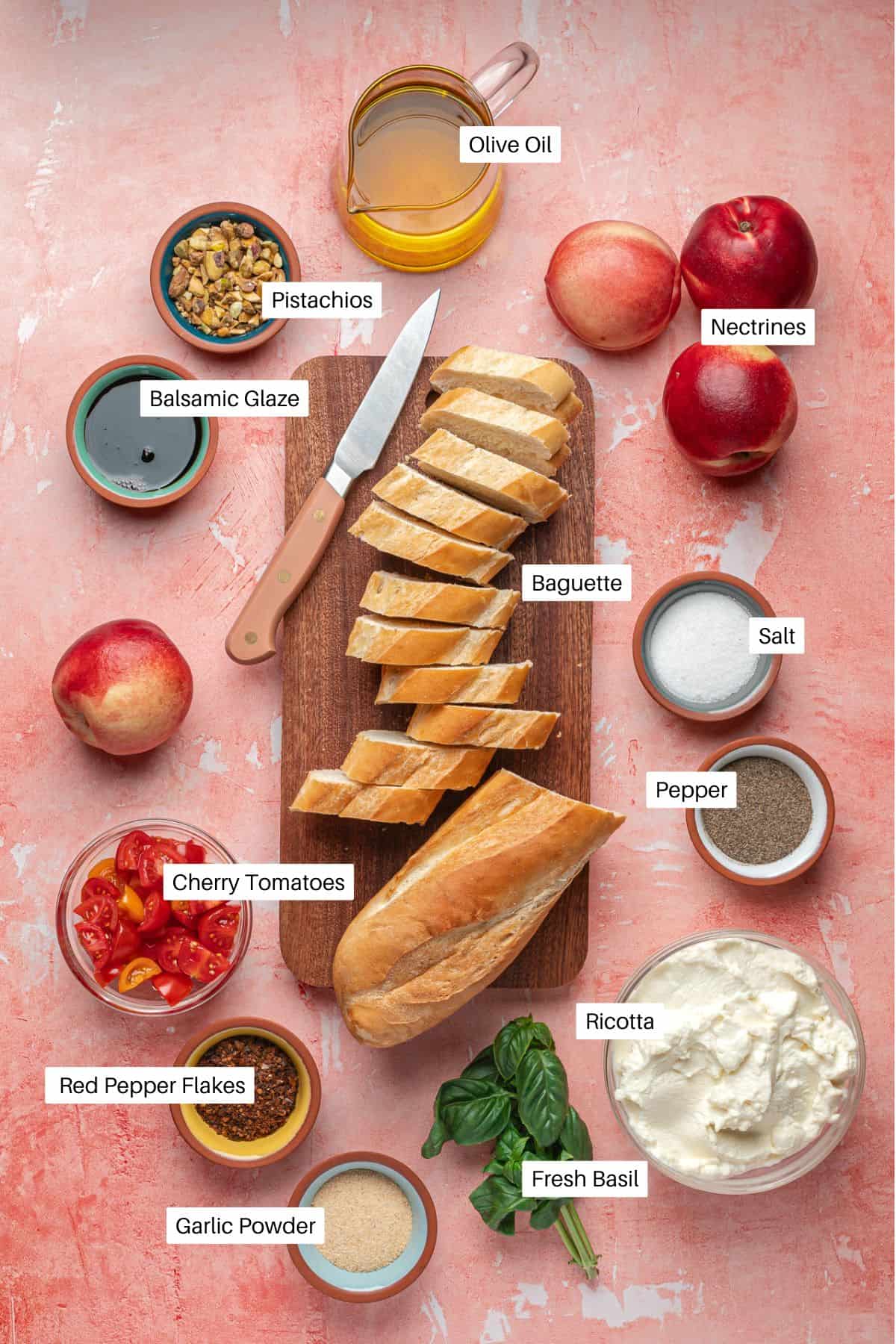 A top-down view of ricotta bruschetta ingredients on a pink surface: sliced baguette, olive oil, pistachios, nectarines, cherry tomatoes, fresh basil, creamy ricotta, salt, pepper, garlic powder, red pepper flakes, and balsamic glaze.