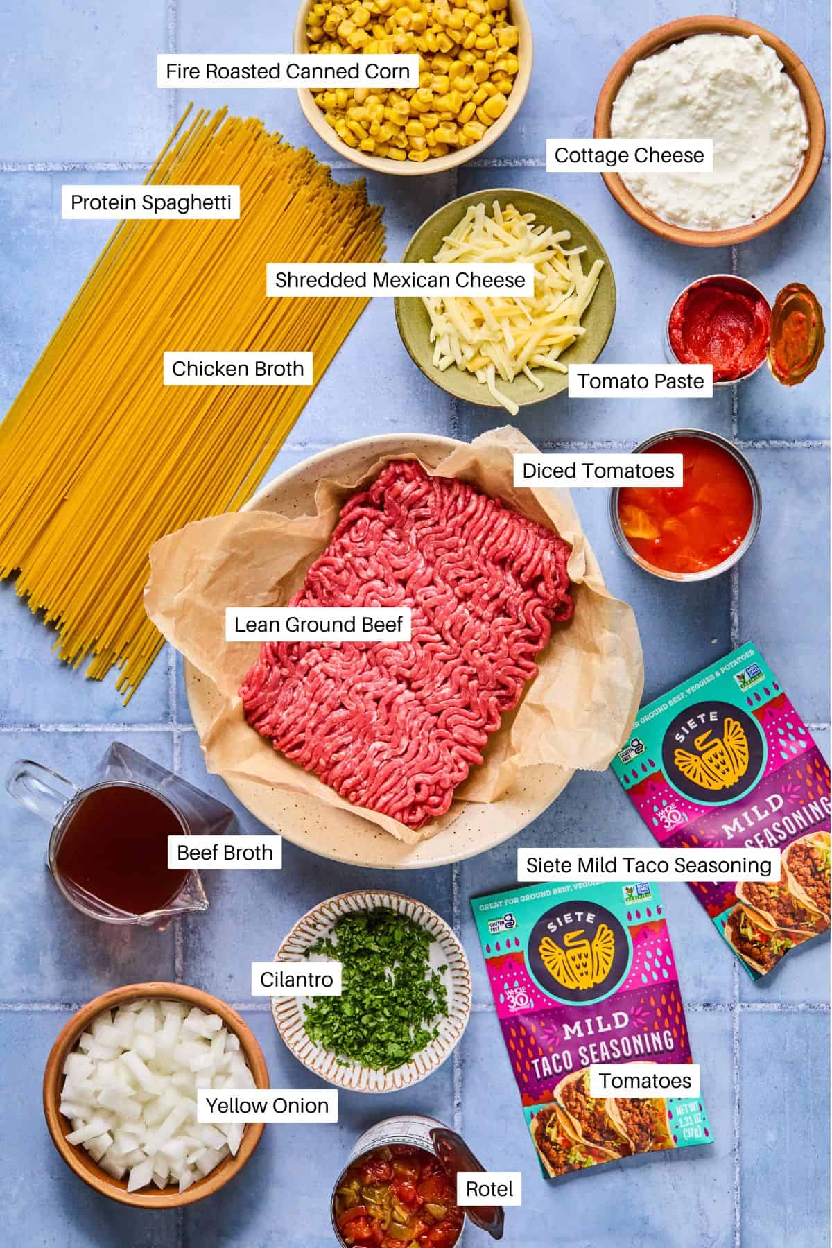 A flat lay of labeled ingredients for taco spaghetti, a Mexican-inspired pasta dish, including ground beef, protein spaghetti, cheeses, broths, canned corn, tomato products, taco seasoning, onion, and chopped cilantro.