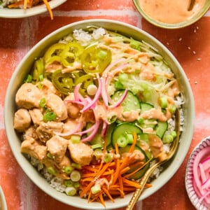 A colorful bowl of rice topped with bang bang chicken, sliced cucumbers, pickled onions, shredded carrots, jalapeños, green onions, and drizzled with a creamy sauce, plus extra sauce in a small bowl nearby.