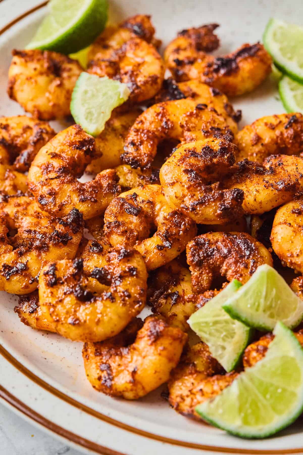 A plate of blackened shrimp garnished with lime wedges, served on a beige dish. The shrimp are golden brown with visible spices, and the limes are sliced into quarters.