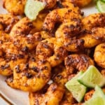 A plate of blackened shrimp garnished with lime wedges, served on a beige dish. The shrimp are golden brown with visible spices, and the limes are sliced into quarters.