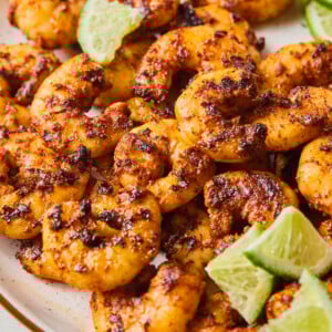 A plate of blackened shrimp garnished with lime wedges, served on a beige dish. The shrimp are golden brown with visible spices, and the limes are sliced into quarters.