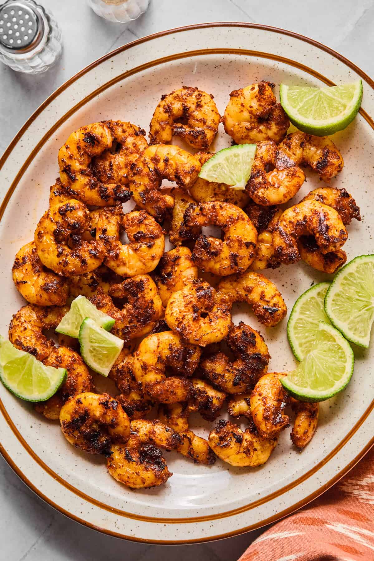 A plate of blackened shrimp garnished with fresh lime wedges, viewed from above. The shrimp are browned and arranged on a white plate with a brown rim, set on a marble surface.