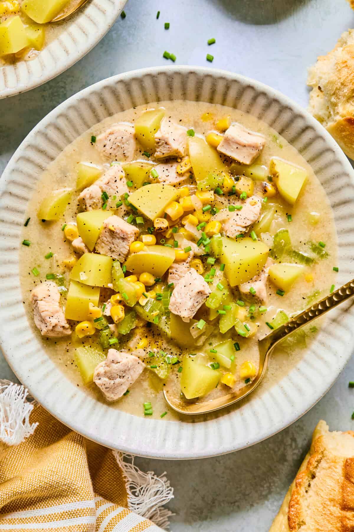A bowl of creamy Corn and Potato Chowder with chicken, corn, chopped chives, and diced celery, served with a gold spoon. Pieces of bread and a yellow napkin are nearby on the table.