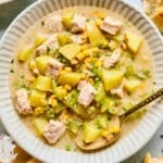 A bowl of creamy Corn and Potato Chowder with chicken, corn, chopped chives, and diced celery, served with a gold spoon. Pieces of bread and a yellow napkin are nearby on the table.