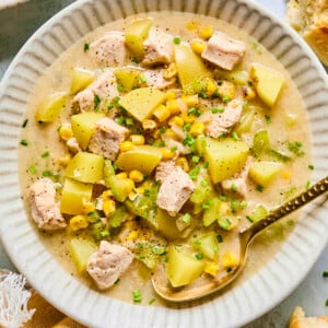 A bowl of creamy Corn and Potato Chowder with chicken, corn, chopped chives, and diced celery, served with a gold spoon. Pieces of bread and a yellow napkin are nearby on the table.