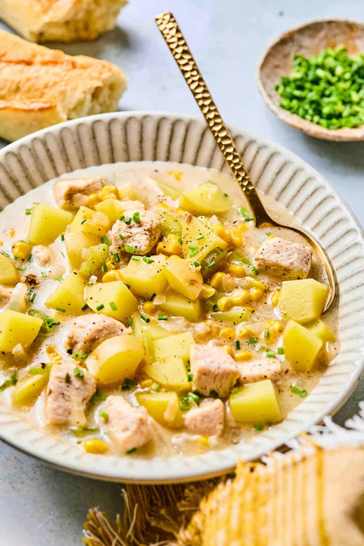 A bowl of creamy Corn and Potato Chowder, garnished with chopped chives. The soup features diced potatoes, tender chicken pieces, and sweet corn. A golden spoon rests in the bowl, with sliced bread and a small dish of chives nearby.