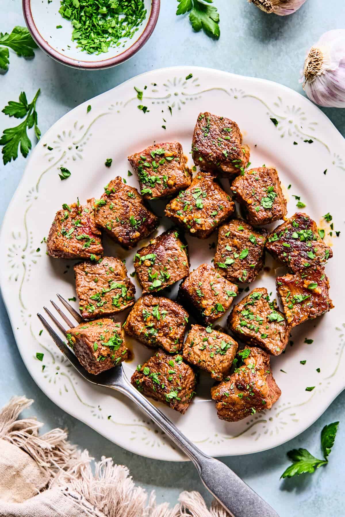 A white plate with garlic steak bites garnished with chopped parsley, served with a fork. Fresh parsley and a bowl of more chopped herbs are nearby on a light blue surface.